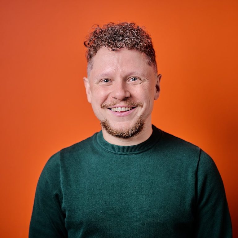 Paul Neal, smiling, with curly hair wearing a green sweatshirt in front of a blue background.